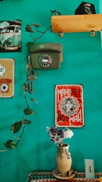 A teal-colored wall is decorated with vintage items, including a rotary dial telephone and a classic poster that reads 'Make Art Not War.' A vine with lush green leaves extends across the wall. Below, a crocheted tablecloth in various colors adorns a surface holding a rustic clay vase filled with a small bouquet of dried flowers. A postcard or card labeled with the number '4' is placed nearby, and a small photographic print of a vintage Volkswagen van is seen in the corner.
