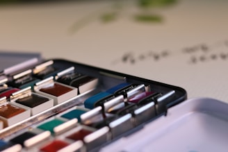 A close-up view of a watercolor paint set featuring multiple colors in small, rectangular pans. The paint set is open, revealing a variety of hues including brown, blue, green, and red. The paints are neatly arranged in a metal box on a white surface with blurred handwritten text in the background.