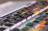 A close-up view of a set of watercolor paints arranged neatly in rows. The paints are packaged in black half-pans with the brand name 'Master Touch' displayed on each. The colors range from vibrant hues like orange, yellow, and green to more subdued shades like purple and blue.