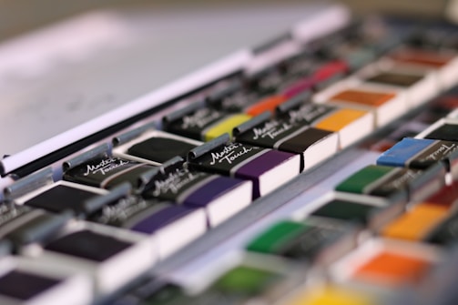 A close-up view of a set of watercolor paints arranged neatly in rows. The paints are packaged in black half-pans with the brand name 'Master Touch' displayed on each. The colors range from vibrant hues like orange, yellow, and green to more subdued shades like purple and blue.