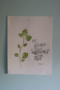 A piece of artwork featuring a simple watercolor painting of a green leafy plant next to an inspirational handwritten quote stating 'My grace is sufficient for thee', attributed to 2 Corinthians 12:9. The painting is on a white paper set against a light blue wall.