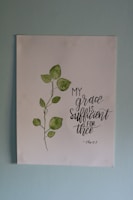 A piece of artwork featuring a simple watercolor painting of a green leafy plant next to an inspirational handwritten quote stating 'My grace is sufficient for thee', attributed to 2 Corinthians 12:9. The painting is on a white paper set against a light blue wall.