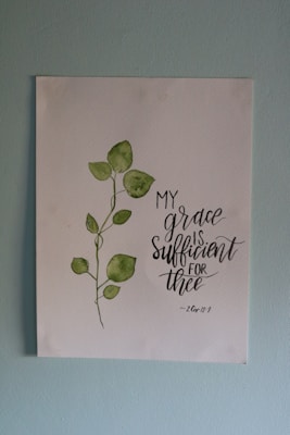 A piece of artwork featuring a simple watercolor painting of a green leafy plant next to an inspirational handwritten quote stating 'My grace is sufficient for thee', attributed to 2 Corinthians 12:9. The painting is on a white paper set against a light blue wall.