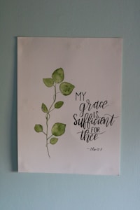 A piece of artwork featuring a simple watercolor painting of a green leafy plant next to an inspirational handwritten quote stating 'My grace is sufficient for thee', attributed to 2 Corinthians 12:9. The painting is on a white paper set against a light blue wall.