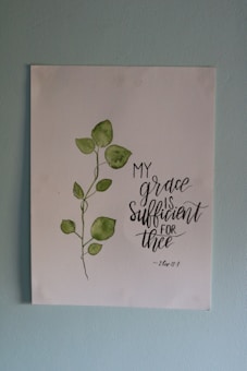 A piece of artwork featuring a simple watercolor painting of a green leafy plant next to an inspirational handwritten quote stating 'My grace is sufficient for thee', attributed to 2 Corinthians 12:9. The painting is on a white paper set against a light blue wall.