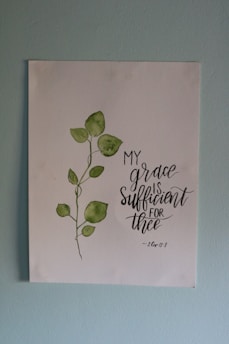 A piece of artwork featuring a simple watercolor painting of a green leafy plant next to an inspirational handwritten quote stating 'My grace is sufficient for thee', attributed to 2 Corinthians 12:9. The painting is on a white paper set against a light blue wall.