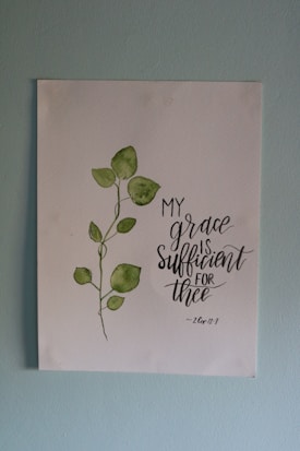 A piece of artwork featuring a simple watercolor painting of a green leafy plant next to an inspirational handwritten quote stating 'My grace is sufficient for thee', attributed to 2 Corinthians 12:9. The painting is on a white paper set against a light blue wall.