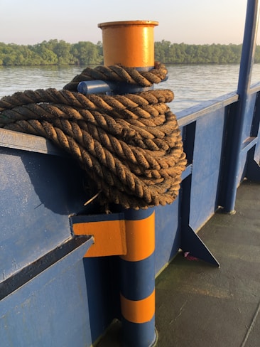 A coiled rope rests on a blue and orange bollard on a boat deck. The background features a view of calm water with a line of lush green trees along the horizon under a clear sky.