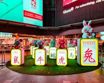 A colorful display featuring cartoonish animal figurines and signs with English and Chinese characters representing different animals of the Chinese zodiac, such as the rat, ox, and rabbit. The figures are placed on illuminated pedestals surrounded by artificial grass, set against the backdrop of a well-lit urban environment with restaurant signage in the background.