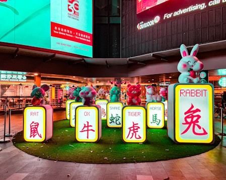 A colorful display featuring cartoonish animal figurines and signs with English and Chinese characters representing different animals of the Chinese zodiac, such as the rat, ox, and rabbit. The figures are placed on illuminated pedestals surrounded by artificial grass, set against the backdrop of a well-lit urban environment with restaurant signage in the background.