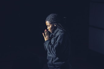 A person with braided hair is shown in a side profile, with hands clasped together in a prayer-like gesture. The individual's expression appears contemplative and serene, surrounded by dim lighting that casts the background in shadow.