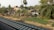 A rural scene featuring a cluster of simple dwellings surrounded by lush greenery and trees. Railway tracks run parallel to the settlement, suggesting connectivity and travel. The area is densely vegetated with banana plants and palm trees, and there's an assortment of makeshift structures.