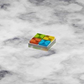 A 3D geometric shape resembling a logo with four colorful squares, each in a different color: orange, green, blue, and yellow. The squares are arranged in a grid on a slightly tilted platform, giving a sense of depth. The background consists of a cloudy pattern in various shades of white and gray.