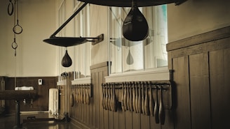 A vintage gym setting with wooden walls and a collection of wooden Indian clubs neatly arranged on a rack. There is an old-school leather speed bag hanging from a hook attached to the wall. Another larger punching bag can be seen on the left side of the room. The room has a nostalgic and historical atmosphere, potentially from an earlier era of fitness.