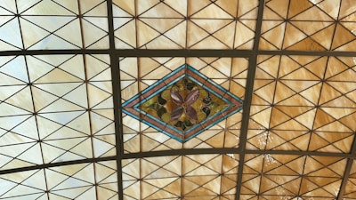 A stained glass ceiling with a diamond-shaped centerpiece featuring a floral design in shades of purple and green. The surrounding glass is arranged in a grid of triangular sections, primarily in shades of cream and amber.