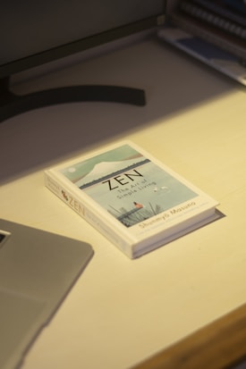 A book titled 'Zen: The Art of Simple Living' rests on a light-colored wooden desk. The cover features minimalist artwork depicting a serene landscape. Nearby, part of a laptop is visible, along with a computer monitor. The setting suggests a calm and organized workspace.