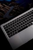 A close-up view of a laptop keyboard and trackpad, with a colorful abstract background peeking underneath the screen. The keyboard features a QWERTY layout with clear lettering, and the laptop casing is metallic with a sleek design.