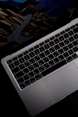 A close-up view of a laptop keyboard and trackpad, with a colorful abstract background peeking underneath the screen. The keyboard features a QWERTY layout with clear lettering, and the laptop casing is metallic with a sleek design.