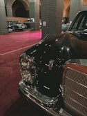 A vintage black car with a chrome grille and circular headlights is prominently displayed in an indoor setting. The space has a maroon carpet and features large, arching columns with intricate patterns. Several other classic cars are visible in the background.