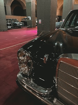 A vintage black car with a chrome grille and circular headlights is prominently displayed in an indoor setting. The space has a maroon carpet and features large, arching columns with intricate patterns. Several other classic cars are visible in the background.