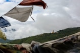 Colorful prayer flags flutter in the foreground with a vast, green forested landscape spread across the background. A large golden statue of a seated figure is visible on a distant hill. The sky is overcast with fluffy white clouds swirling over the treetops.
