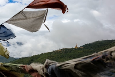 Colorful prayer flags flutter in the foreground with a vast, green forested landscape spread across the background. A large golden statue of a seated figure is visible on a distant hill. The sky is overcast with fluffy white clouds swirling over the treetops.
