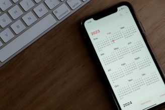 A smartphone displaying a 2023 yearly calendar on its screen is placed on a wooden surface. To the left, part of a white computer keyboard with keys including control, option, and command is visible.
