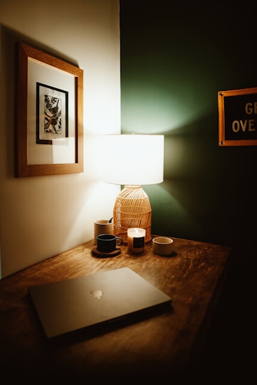 A cozy and dimly lit corner of a room features a wooden desk with a laptop, a lit lamp with a woven shade, a framed artwork on the wall, a couple of candles, and a black mug on a saucer. The wall has a sign partially visible with text.