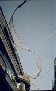 A large, curved net-like structure is suspended between two buildings against a clear blue sky. The buildings are modern with reflective glass windows and sleek architectural details. The installation appears to be an art piece or architectural feature.