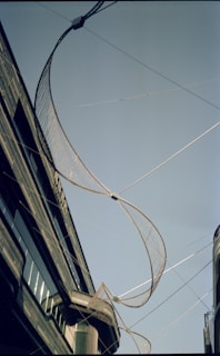 A large, curved net-like structure is suspended between two buildings against a clear blue sky. The buildings are modern with reflective glass windows and sleek architectural details. The installation appears to be an art piece or architectural feature.