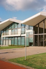 A modern building with extensive glass windows and a uniquely designed roof. The facade features large glass panels and balconies. In front of the building, there's a directional signpost situated on a well-maintained lawn, and a paved pathway leads up to the entrance. The sky above is partly cloudy with patches of blue visible.