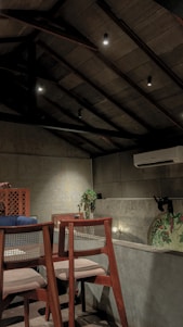 A cozy interior setting featuring wooden chairs with woven backs, situated next to a low concrete wall. A potted plant adds a touch of greenery, while an air conditioning unit is visible on the wall. The warm lighting from the overhead fixtures creates a welcoming ambiance, complementing the detailed mural that can be partially seen.