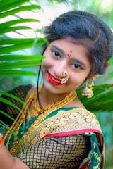 A woman is adorned in traditional attire, featuring a richly designed green sari with gold patterns. She wears elaborate jewelry, including a nose ring and earrings, and has her hair styled neatly. She is smiling with bright red lipstick, surrounded by green leaves, suggesting a natural or garden setting.