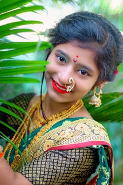 A woman is adorned in traditional attire, featuring a richly designed green sari with gold patterns. She wears elaborate jewelry, including a nose ring and earrings, and has her hair styled neatly. She is smiling with bright red lipstick, surrounded by green leaves, suggesting a natural or garden setting.