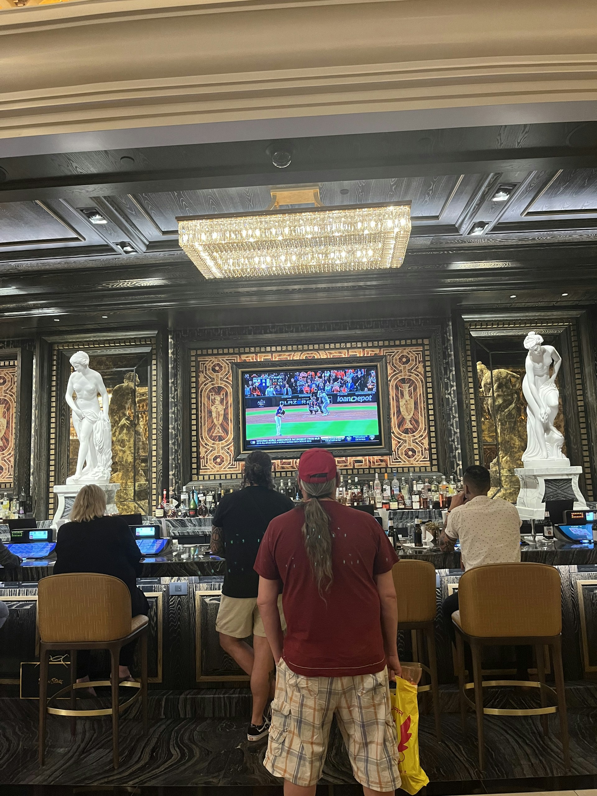 Several people are seated at a luxurious bar, with a large TV screen showing a sports game. The bar is elegantly decorated with ornate details and sculptures. The ceiling has a large, rectangular, crystal chandelier.