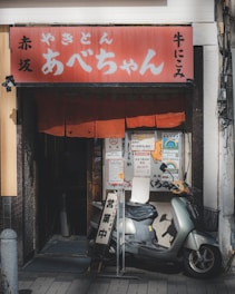 A small street-side restaurant has a traditional red sign with Japanese writing above the entrance, and a red noren curtain hanging in front. A silver scooter is parked outside next to standing signboards filled with Japanese text and promotional flyers.