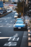 A city street scene featuring a blue sports car parked on the side of a busy road with visible Japanese characters painted on the pavement. Other vehicles, including a white van, are driving on the road. Trees line the sidewalk, and there are various signs and buildings in the background.