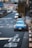 A city street scene featuring a blue sports car parked on the side of a busy road with visible Japanese characters painted on the pavement. Other vehicles, including a white van, are driving on the road. Trees line the sidewalk, and there are various signs and buildings in the background.