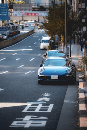 A city street scene featuring a blue sports car parked on the side of a busy road with visible Japanese characters painted on the pavement. Other vehicles, including a white van, are driving on the road. Trees line the sidewalk, and there are various signs and buildings in the background.