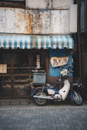 A vintage motorcycle is parked in front of a quaint shop with a faded, weathered exterior. The shop has a blue and white striped awning and signs with Japanese lettering are displayed. The motorcycle has a box on the back, suggesting it's used for deliveries.