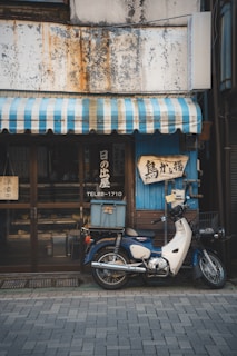 A vintage motorcycle is parked in front of a quaint shop with a faded, weathered exterior. The shop has a blue and white striped awning and signs with Japanese lettering are displayed. The motorcycle has a box on the back, suggesting it's used for deliveries.