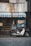 A vintage motorcycle is parked in front of a quaint shop with a faded, weathered exterior. The shop has a blue and white striped awning and signs with Japanese lettering are displayed. The motorcycle has a box on the back, suggesting it's used for deliveries.