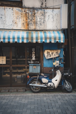 A vintage motorcycle is parked in front of a quaint shop with a faded, weathered exterior. The shop has a blue and white striped awning and signs with Japanese lettering are displayed. The motorcycle has a box on the back, suggesting it's used for deliveries.