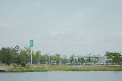A gas station is located in a spacious area with a clear blue sky overhead. The foreground features a body of water bordered by greenery. In the background, the station has a sleek design with a prominent teal and white sign. Cars are parked and people are visible moving around, while trees and bushes add a natural touch to the scene.