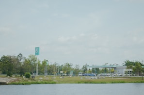 A gas station is located in a spacious area with a clear blue sky overhead. The foreground features a body of water bordered by greenery. In the background, the station has a sleek design with a prominent teal and white sign. Cars are parked and people are visible moving around, while trees and bushes add a natural touch to the scene.