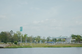 A gas station is located in a spacious area with a clear blue sky overhead. The foreground features a body of water bordered by greenery. In the background, the station has a sleek design with a prominent teal and white sign. Cars are parked and people are visible moving around, while trees and bushes add a natural touch to the scene.