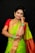 A woman in traditional attire, adorned with elaborate gold jewelry and a vibrant saree. Her hair is neatly styled and she has henna designs on her hand. The saree features a combination of green and red colors with intricate patterns.