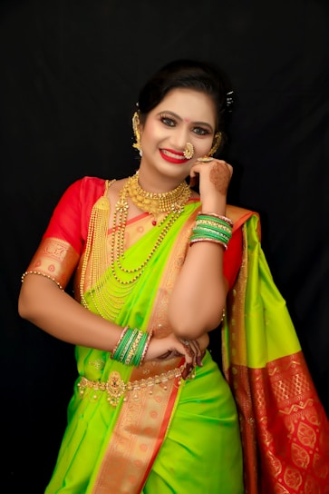 A woman in traditional attire, adorned with elaborate gold jewelry and a vibrant saree. Her hair is neatly styled and she has henna designs on her hand. The saree features a combination of green and red colors with intricate patterns.