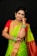 A woman in traditional attire, adorned with elaborate gold jewelry and a vibrant saree. Her hair is neatly styled and she has henna designs on her hand. The saree features a combination of green and red colors with intricate patterns.