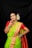 A woman dressed in a traditional green and orange saree adorned with gold jewelry, including necklaces, earrings, and a nose ring. She has her hair styled with white flowers and is wearing colorful bangles on her wrists. Her pose is confident and she is smiling.